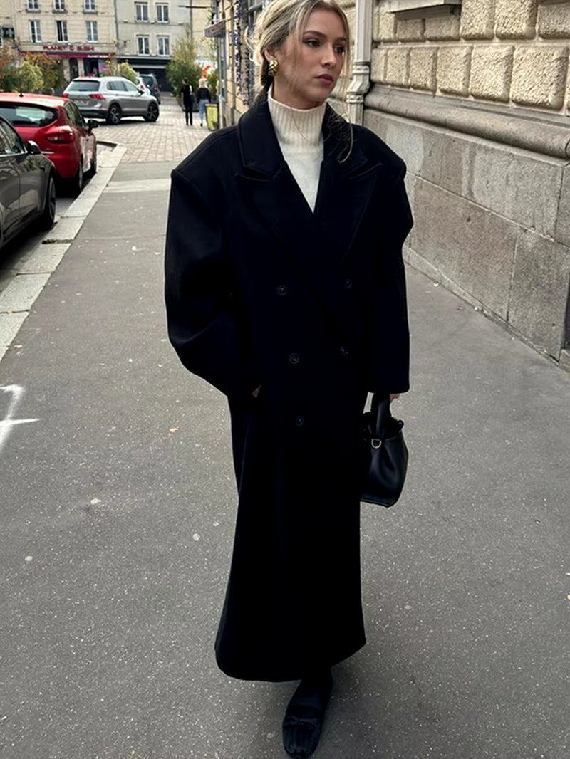 Model draagt zwarte Vesper Luxe Coat met handtas en laarzen op straat
