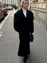 Model draagt zwarte Vesper Luxe Coat met handtas en laarzen op straat
