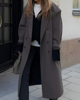 Model in Vesper Luxe Coat bruin voor een zwarte deur, met stijlvolle accessoires
