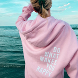 Frau trägt pastellrosa Hoodie mit Textdruck "Do What Makes You Happy" am Meer