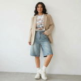 Modell trägt Vayla Denim Bermuda Shorts in Hellblau mit beigem Oversized-Blazer, Graphic Crop Top und weißen Sneakers – Streetstyle Look